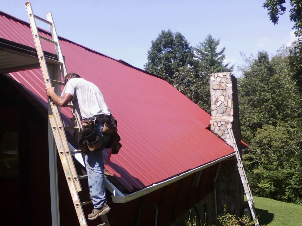 Roofer on a ladder beside a red metal roof and stone chimney — Soffit & Fascia Repair by Roofing Brothers in Port Matilda